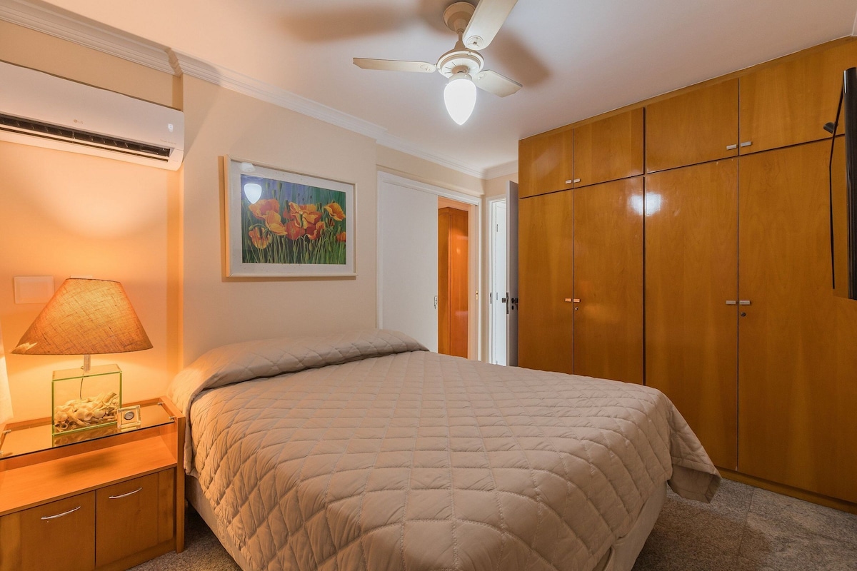 Ein Schlafzimmer mit einem Bett, einem Nachttisch, einer Klimaanlage, einem Deckenventilator, Holzschränken und einem gerahmten Bild an der Wand.