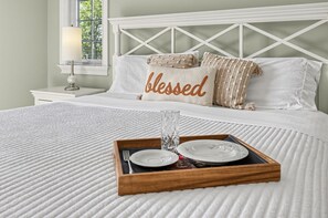 Ein ordentlich bezogenes Bett mit einem Tablett, darunter Teller und ein Glas, einer Nachttischlampe sowie Kissen mit dem Aufdruck 'blessed'.