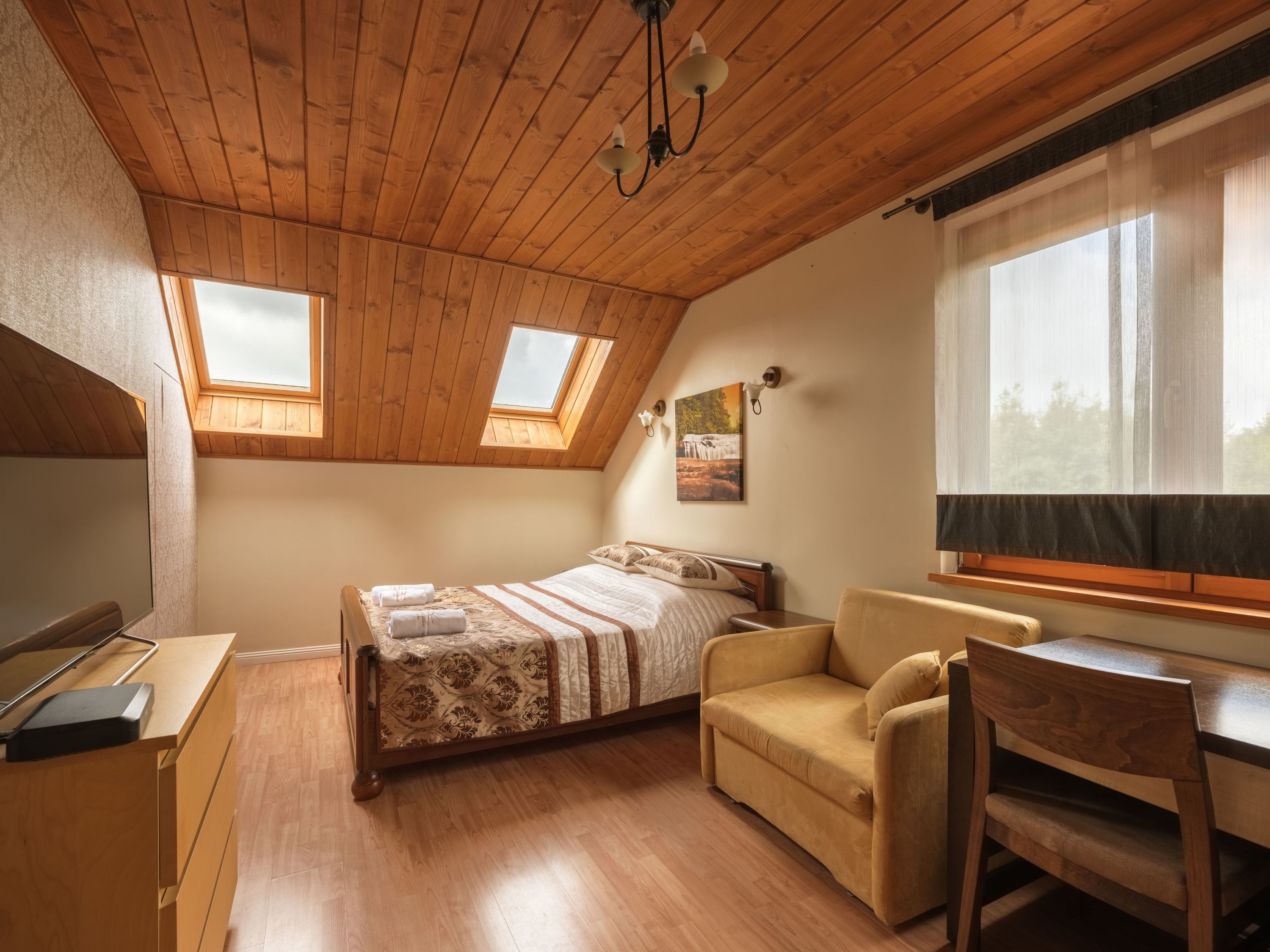 Un dormitorio con techo inclinado, vigas de madera, una cama, un sofá, un escritorio y dos tragaluces.