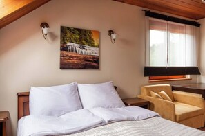 Ein Schlafzimmer mit einem Bett, einem Sessel, einem Nachttisch, einer Lampe, einem Bild eines Wasserfalls und einem Fenster mit Vorhängen.