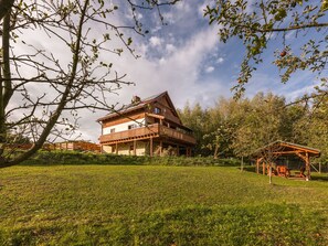 A two-story wooden house with a balcony, surrounded by a grassy area and trees.