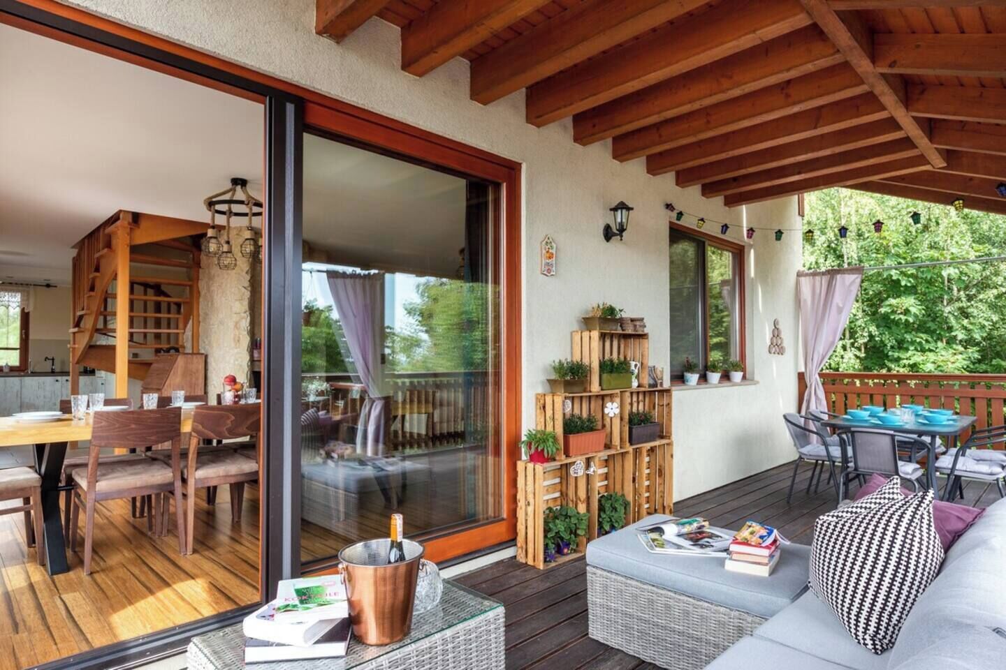 Een overdekt buitengebied met een houten terras, een eettafel met stoelen, een bank en een kleine tafel met een fles en een menukaart.