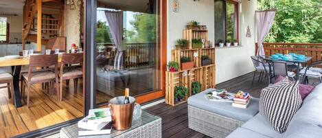 Un espacio al aire libre techado con terraza de madera, una mesa de comedor con sillas, un sofá y una mesita con una botella y un menú.