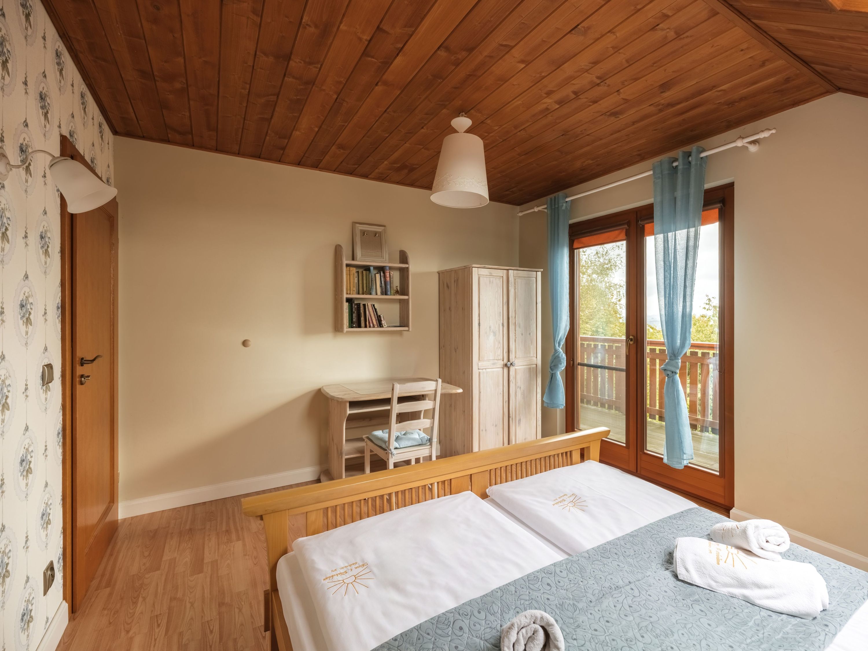 Een slaapkamer met een houten plafond, een bed met witte lakens, een houten bureau, een kledingkast en een raam met gordijnen.