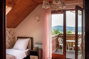 Une chambre avec un plafond en bois, un lit avec une literie blanche, une table de chevet, une fenêtre donnant sur un paysage verdoyant et des montagnes, et un balcon avec des chaises et une table blanches.