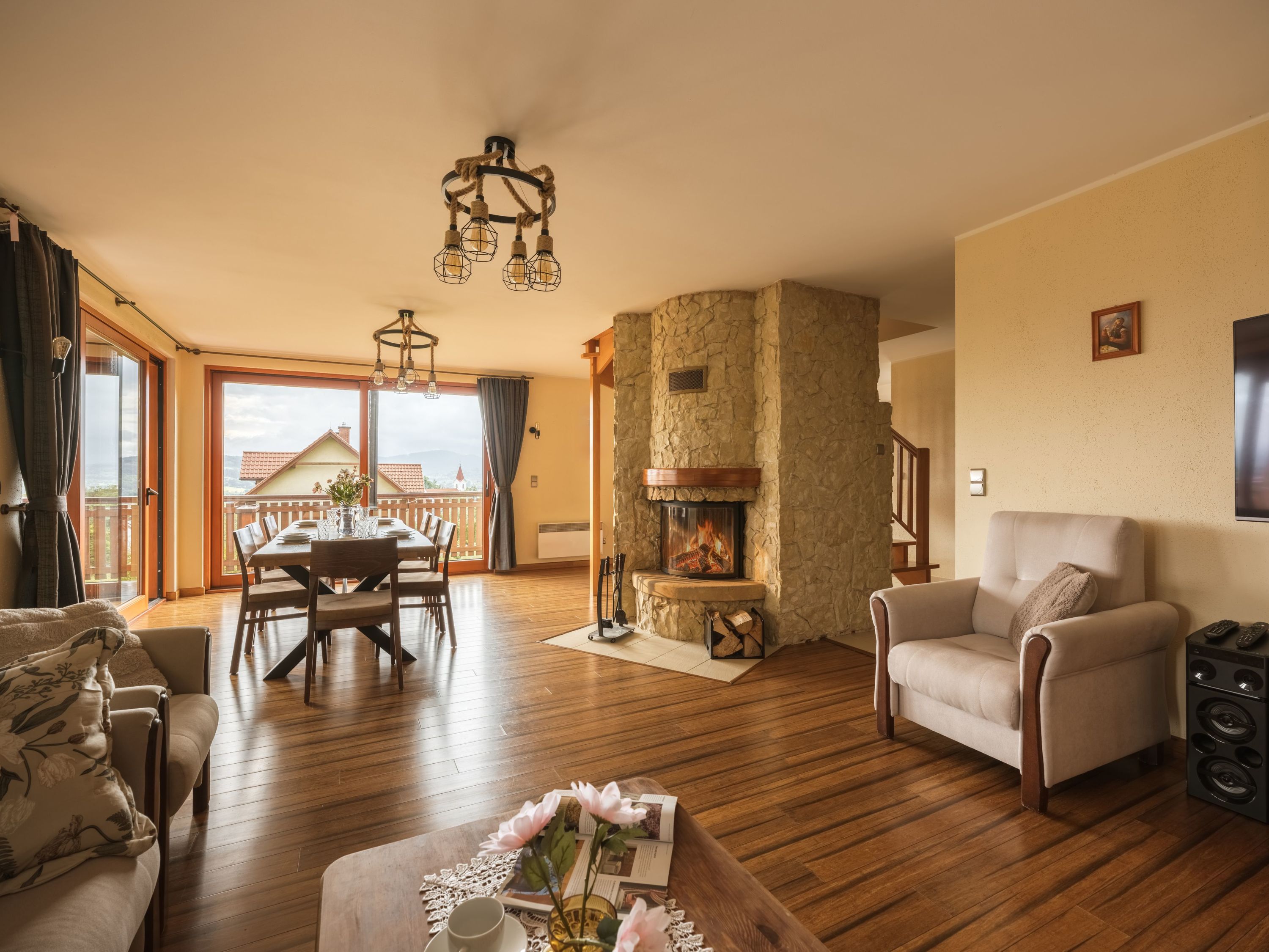 A spacious living room with a fireplace, wooden flooring, a dining area, and a balcony with a view.