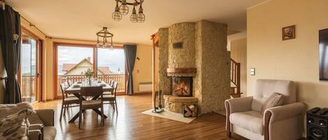 A spacious living room with a fireplace, wooden flooring, a dining area, and a balcony with a view.