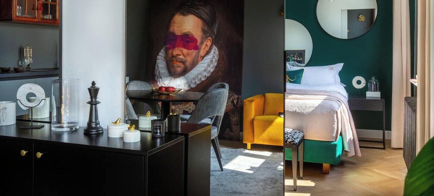 A hotel room with a dining area, a large painting, a bed with a green headboard, and a round mirror.