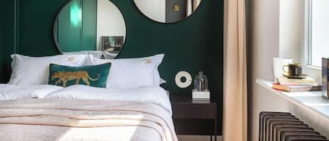 A neatly made bed with white linens and a leopard print pillow, a bedside table with a lamp and books, and a window with curtains.
