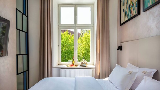 Un dormitorio con un ventanal grande, una cama con ropa de cama blanca, un radiador y vistas a la vegetación exterior.