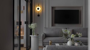 Ein modernes Wohnzimmer mit einem grauen Sofa, einem runden Tisch mit Blumenvase, einem an der Wand montierten Fernseher und einer stehenden Stehlampe.