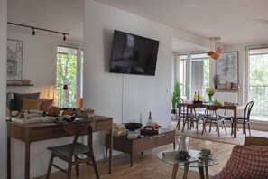 A modern living room with a flat-screen TV, wooden furniture, a dining area with a table and chairs, and large windows allowing natural light. - Modern 2-Bed Apartment with 2 Bathrooms, Balcony (Kraków)