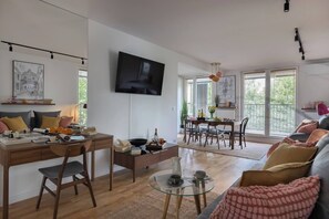 A modern living room with a dining area, a flat-screen TV mounted on the wall, and a large window providing natural light. - Modern 2-Bed Apartment with 2 Bathrooms, Balcony (Kraków)