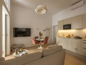 Un salon moderne comprenant un canapé, une table à manger avec des chaises, un téléviseur fixé au mur et un coin cuisine avec des placards et des appareils électroménagers.