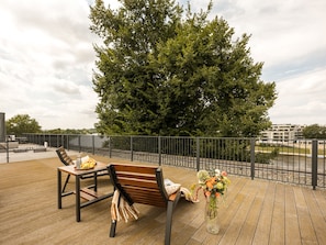 A wooden deck with outdoor furniture, a metal fence, and a large tree in the background. - Vistula View Roof terrace 1-bed Apartment (Kraków)