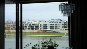 Ein modernes Gebäude mit mehreren Etagen, ein Fluss im Vordergrund und eine Ansicht durch ein Fenster mit einem Topf auf der Fensterbank.