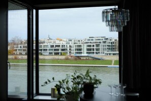 A modern building with multiple floors, a river in the foreground, and a view through a window with a plant on the windowsill. - Vistula View 1-bed Apartment (Kraków)
