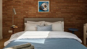 Un dormitorio con pared de ladrillo, cama con ropa de cama blanca, mesita de noche con lámpara y un cuadro sobre la cama.