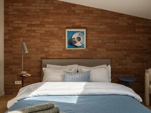Ein Schlafzimmer mit Backsteinwand, einem Bett mit weißer Bettwäsche, einem Nachttisch mit Lampe und einem Gemälde über dem Bett.