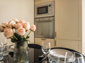 A dining area with a table set for two, a vase of flowers, and a kitchen with a microwave and oven. - White Lotus - 2-bed apartment, full kitchen (Kraków)