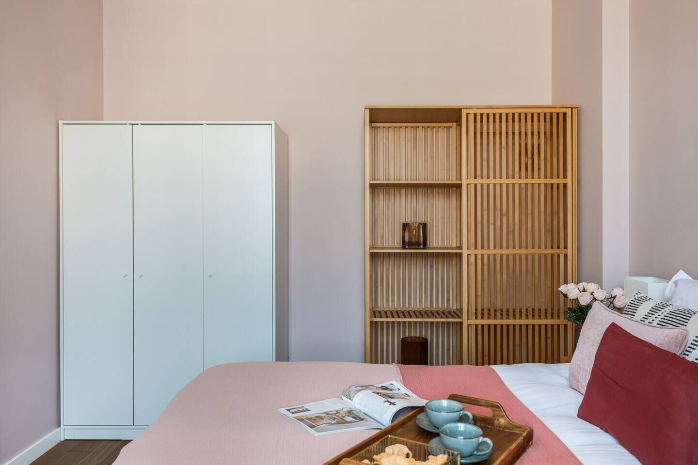 A bedroom with a bed, a wardrobe, a wooden shelf unit, and a tray with items on the bed.