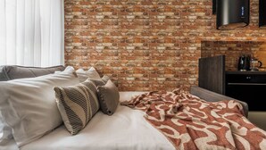 Ein Hotelzimmer mit Backsteinwand, einem Bett mit weißer Bettwäsche und gemusterten Kissen, einer Decke, einem dunklen Holz-Kopfteil, einem flachbildfernseher an der Wand und einer kleinen Küchenzeile mit Kaffeemaschine.