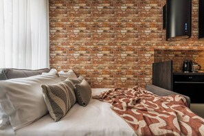 A hotel room with a brick wall, a bed with white bedding and patterned pillows, a blanket, a dark wooden headboard, a flat-screen TV mounted on the wall, and a small kitchen area with a coffee maker. - Skawińska 2-bed apartment on the ground floor (Kraków)