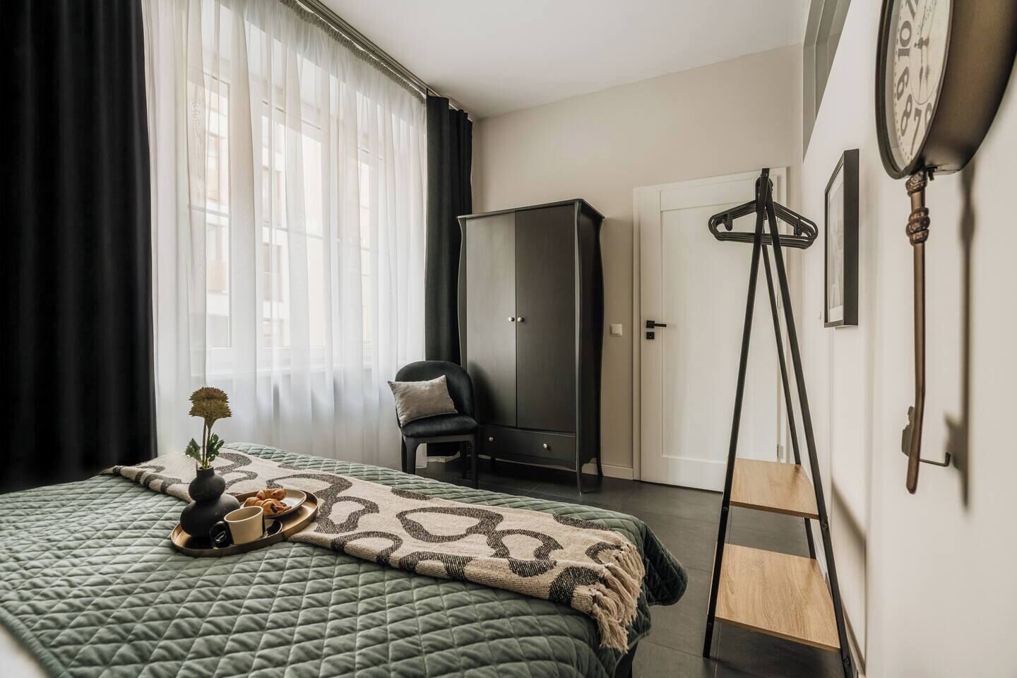A bedroom with a bed, a wardrobe, a chair, a clock, and a small table with a vase.