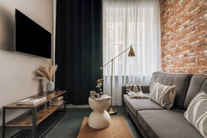A modern living room with a sofa, a coffee table, a TV mounted on the wall, a brick wall, and a window with sheer curtains. - Skawińska 2-bed apartment on the ground floor (Kraków)