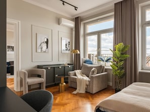 A modern hotel room with a sofa, armchair, and a bed. There is a large window with curtains, a potted plant, and artwork on the wall. - Iconic Sunset Penthouse 115 sqm (Kraków)