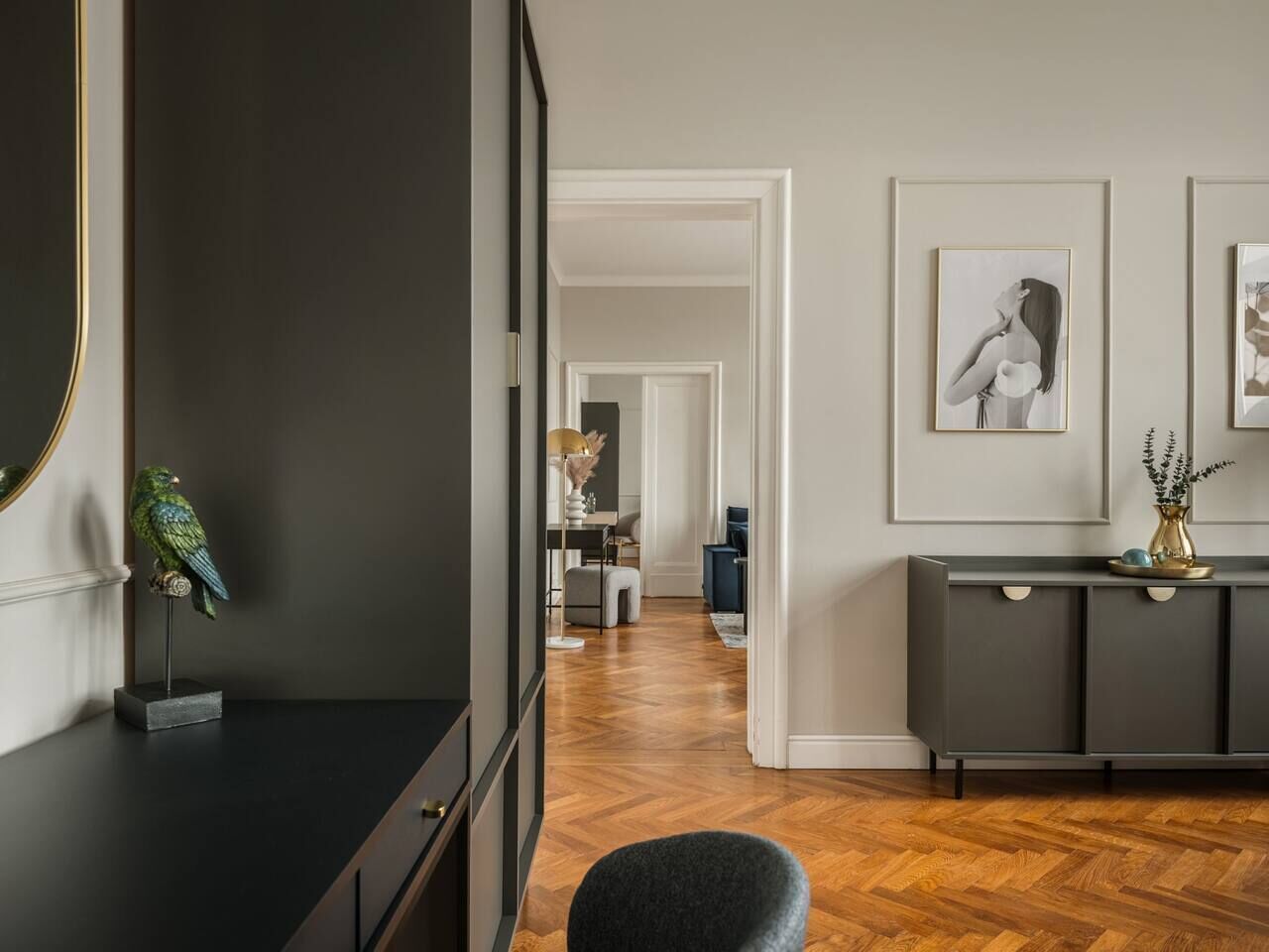 Un corridoio con una parete scura, pavimento in legno, un mobile contenitore grigio con un vaso e quadri appesi al muro.