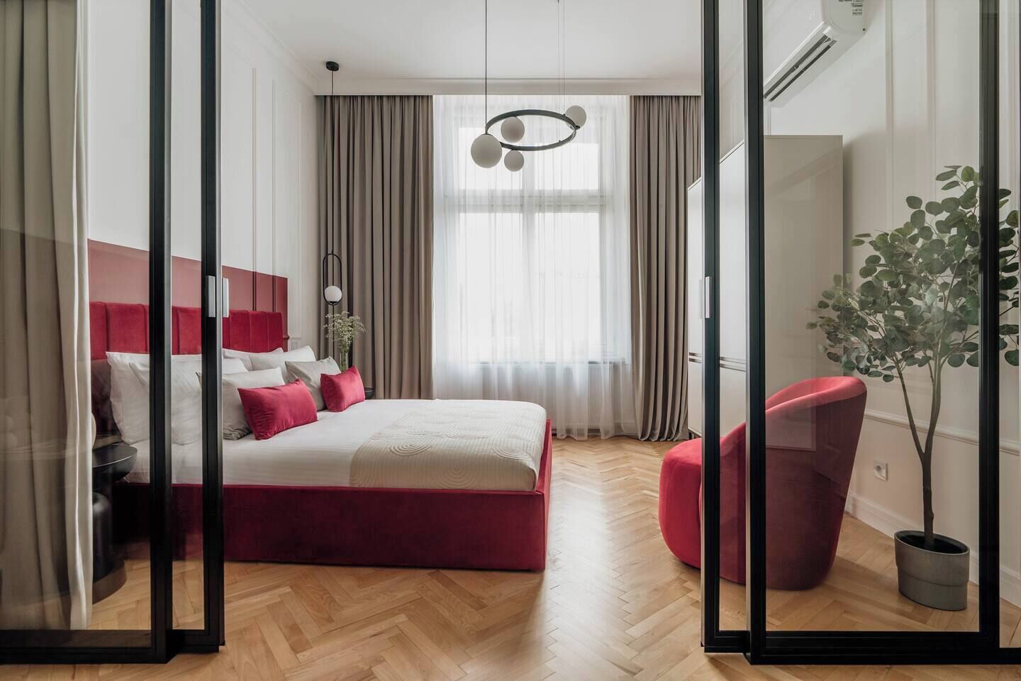 Une chambre à coucher moderne avec un lit, un fauteuil rouge, une plante en pot et une grande fenêtre avec des rideaux.