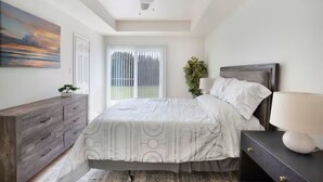 Quarto com cama, penteadeira, mesa de cabeceira, ventilador de teto, janela com persianas e um quadro na parede.