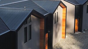 En række moderne hytter med skifertage, beliggende på en grusvej, med et bjergrigt landskab og et vandområde i horisonten.