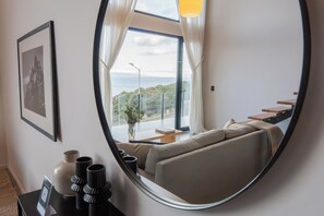 Ein modernes Wohnzimmer mit einem großen Spiegel, der einen Blick auf einen Balkon reflektiert, einer Couch, einem kleinen Tisch und einem gerahmten Bild an der Wand.