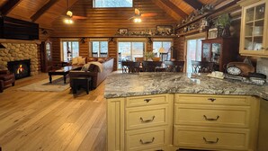 A log cabin interior with a stone fireplace, wooden ceiling, and a dining area with a large island counter.