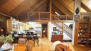 A spacious living area with a wooden ceiling, a dining table with chairs, a brown leather sofa, a staircase, and a bookshelf.