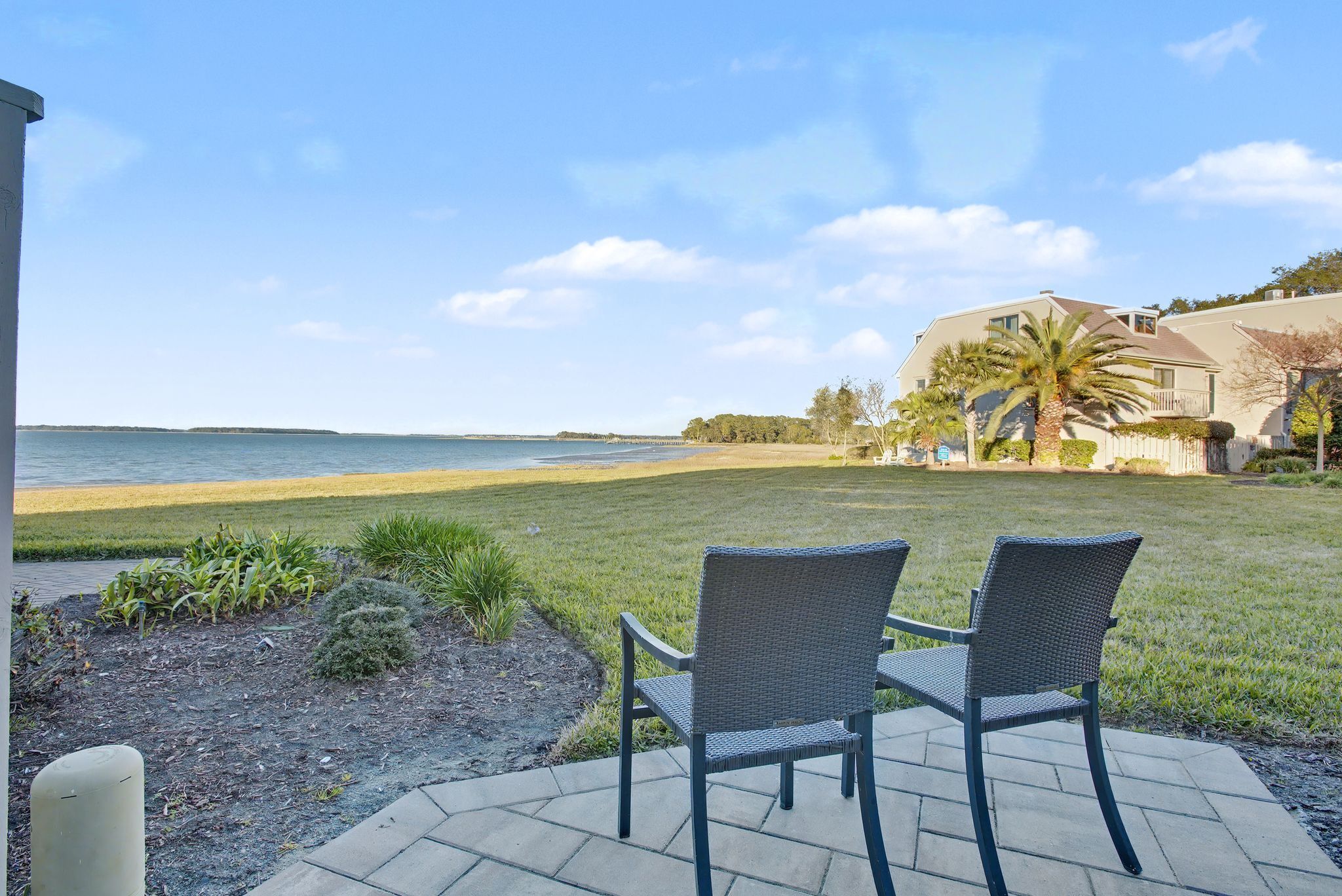 A patio with two chairs facing a beach, a lawn, and a house in the background.