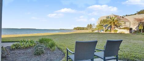 A patio with two chairs facing a beach, a lawn, and a house in the background.