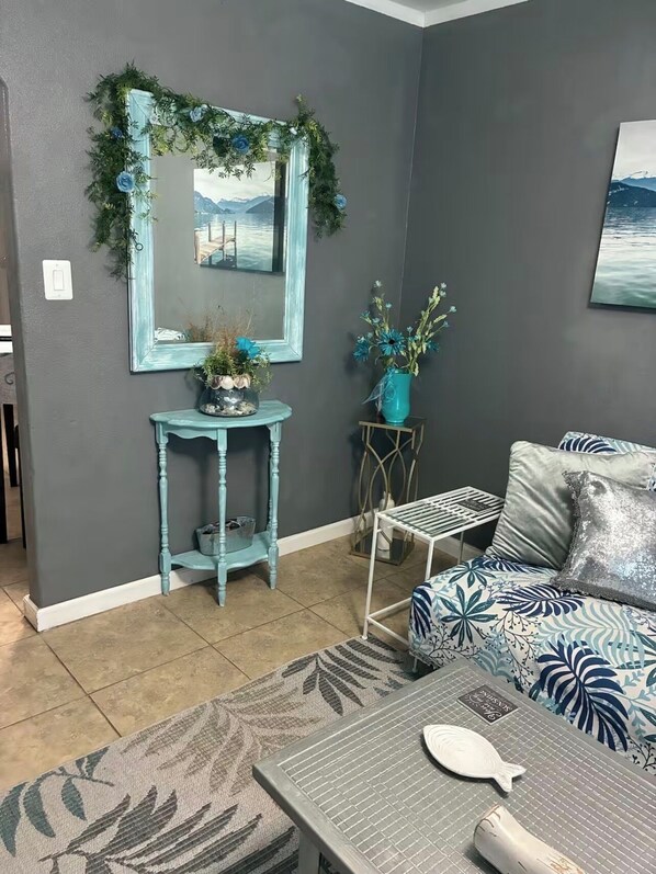 A living room with a grey wall, a mirror with a blue frame, a blue sofa with patterned cushions, a small table with a vase, and a rug with a leaf pattern.