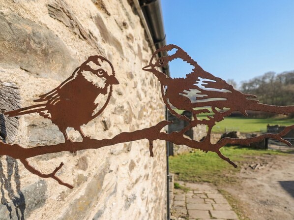 Une découpe métallique représentant deux oiseaux perchés sur une branche, le tout placé devant un mur de pierre.
