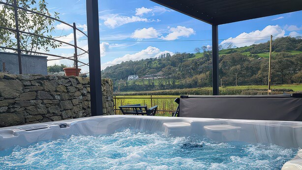 A hot tub with a view of a green landscape and hills in the background. - The Old Cartshed Yr Hen Gwt Cart (Abergele)