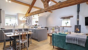 A spacious loft with a high ceiling, exposed wooden beams, a kitchen with a large clock, a dining area with a wooden table and chairs, a cozy living area with a sofa, and a fireplace. - The Old Cartshed Yr Hen Gwt Cart (Abergele)