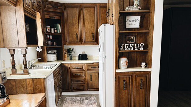 A kitchen with wooden cabinets, a white refrigerator, a wooden dining table, and a patterned rug. - Cozy 2-Bedroom Hulett Home Near Devils Tower & Black Hills (Hulett)