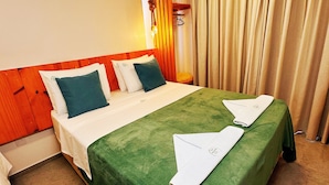 A hotel room with a large bed, a smaller bed, and a green bedspread. There is a wooden headboard, a hanging light fixture, and a window with curtains.