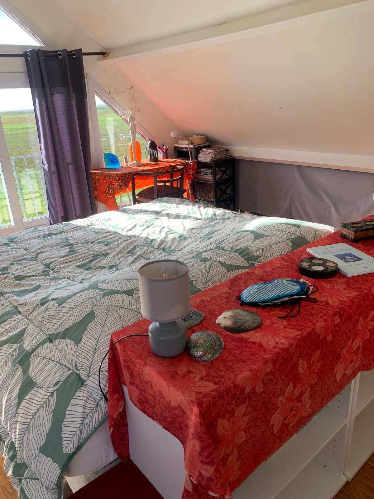 Ein Schlafzimmer mit einem Bett, einem Schreibtisch und einem Stuhl. Es gibt ein Fenster mit Vorhängen, eine Lampe und verschiedene Gegenstände auf dem Schreibtisch.