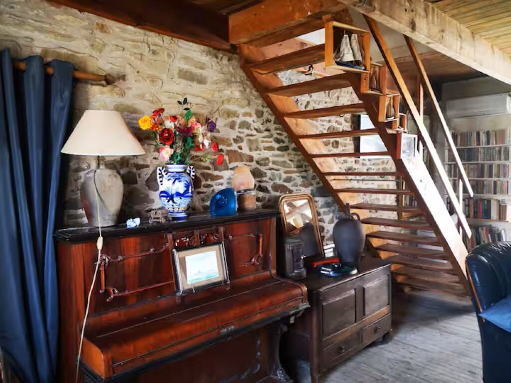 A rustic room with a stone wall, wooden staircase, a wooden dresser, a lamp, and a vase with flowers.