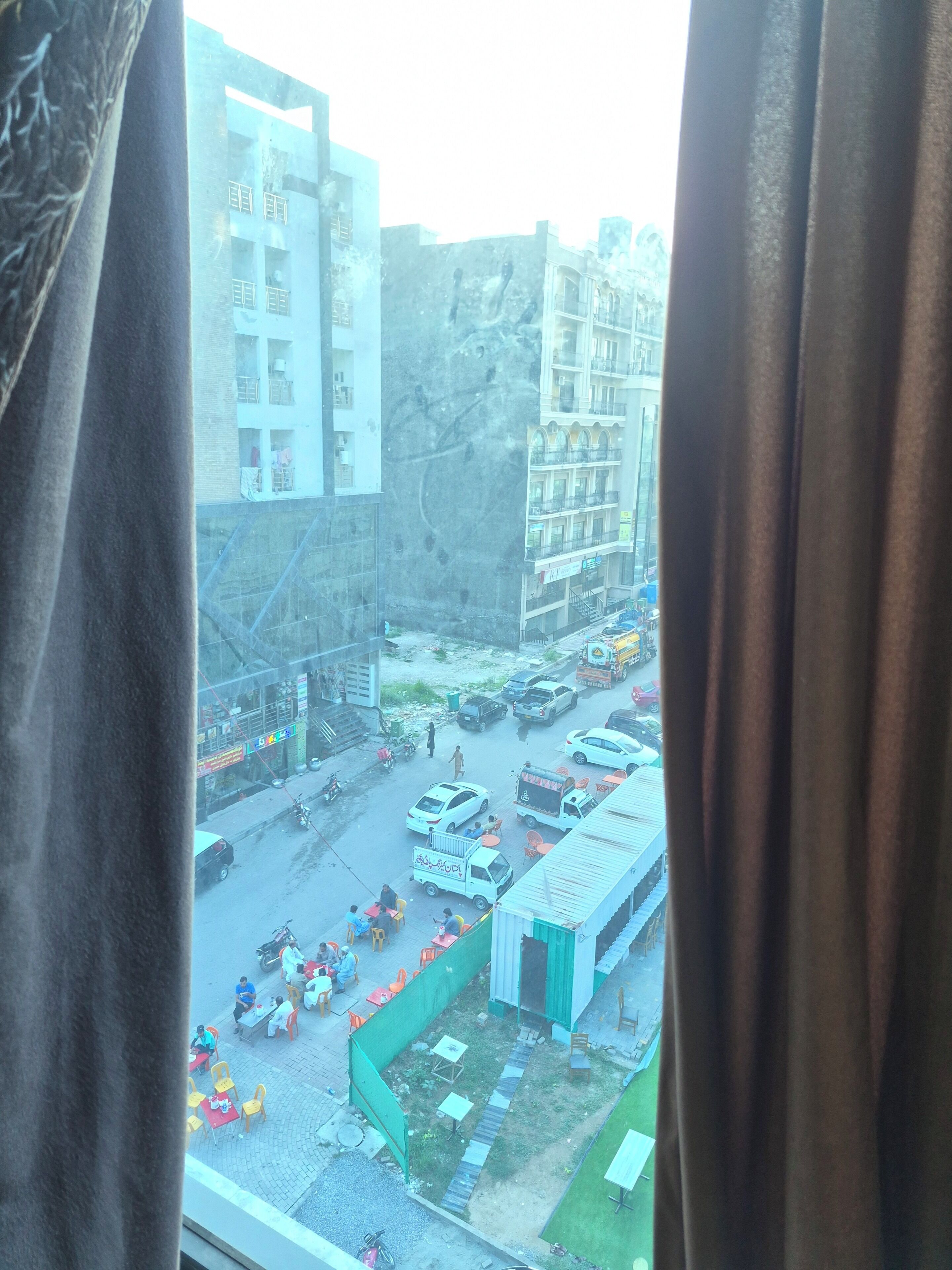 Une vue d’une rue animée, avec des immeubles, une clôture verte et une voiture blanche garée.