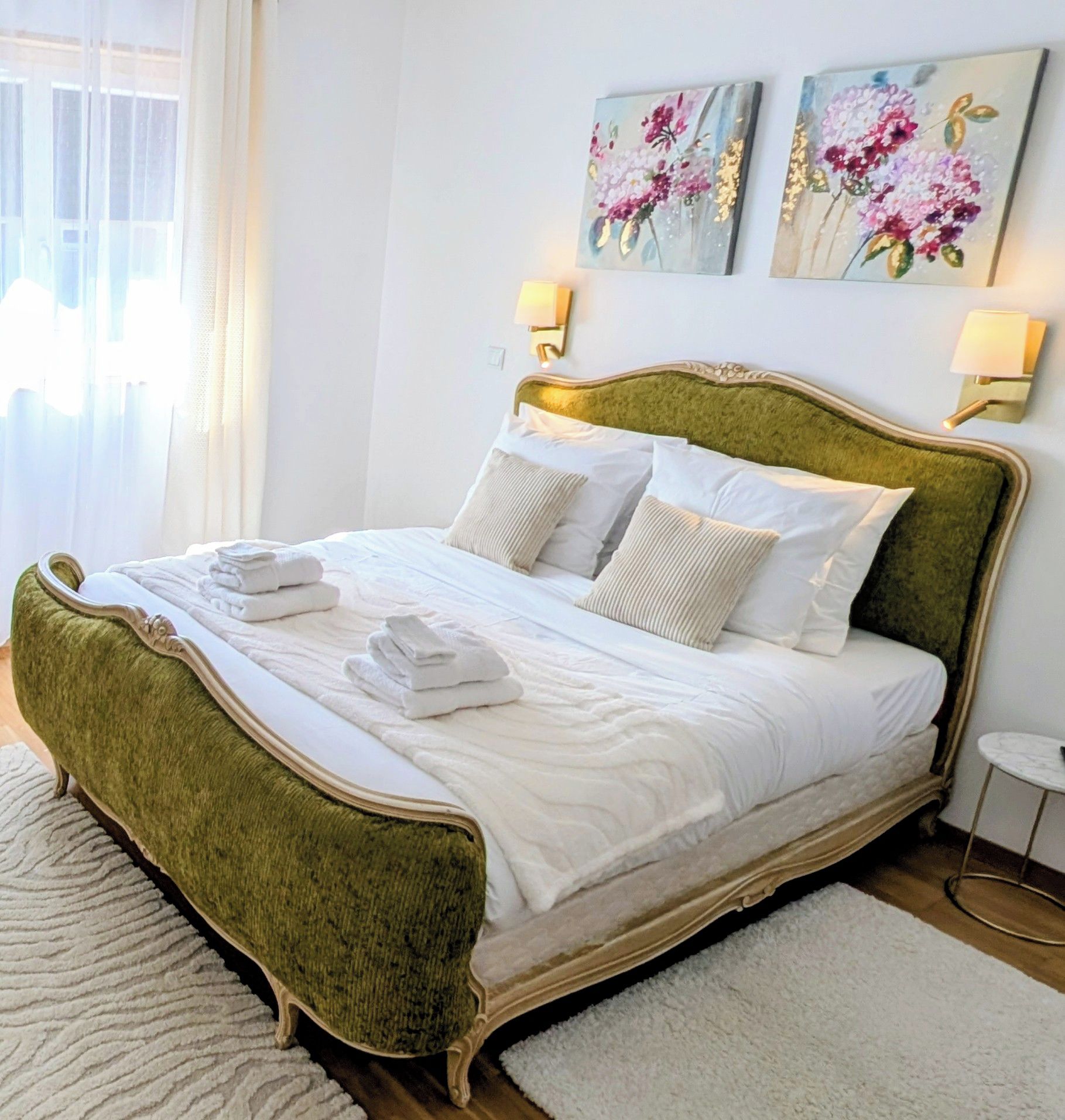Un lit avec une tête de lit rembourrée de tissu vert, une literie blanche et deux lampes fixées au mur.
