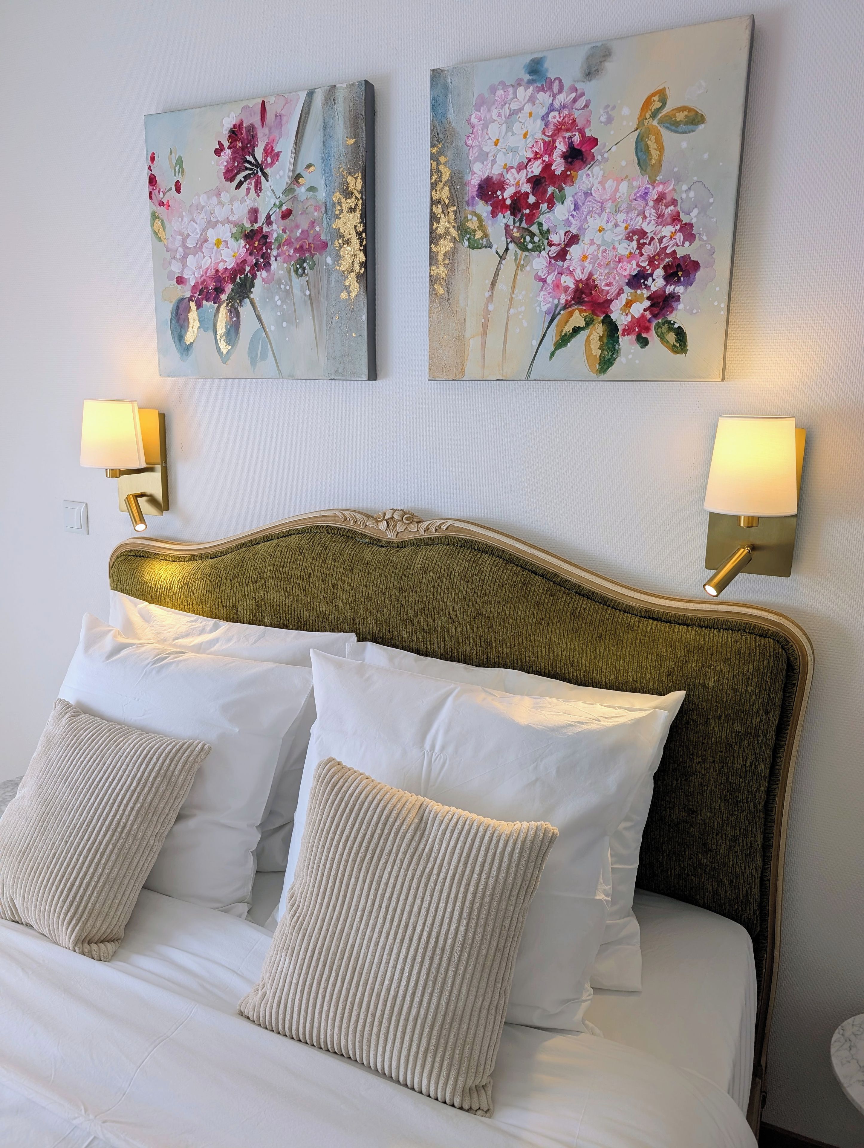 A neatly made bed with white linens and beige pillows, flanked by two wall-mounted lamps with white shades.